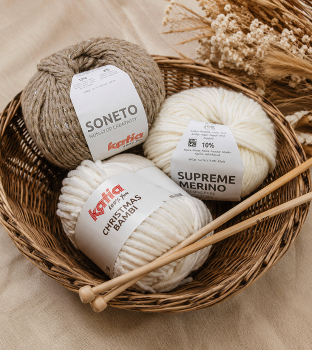 Pelotes de laine Katia Soneto, Supreme Merino et Christmas Bambi présentées dans un panier en osier avec des aiguilles à tricoter en bois sur un tissu naturel.